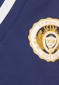 Camisa de algodón azul con un logo gráfico redondo en amarillo y blanco que presenta una corona y hojas, ubicado en el lado izquierdo cerca del escote.