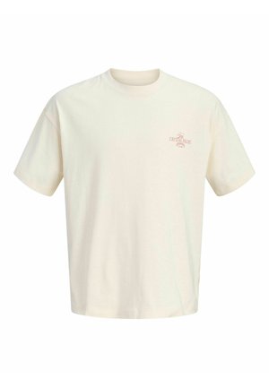 Chłopiec stoi, ubrany w kremowy T-shirt, ciemne dżinsowe szorty, kremowe trampki, sportowe skarpetki oraz czerwoną czapkę z daszkiem na jednolitym tle.
