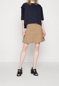 Donna che indossa una maglia nera larga, una gonna beige arricciata e sandali neri con tacco a blocco aperti davanti, dettagli in oro, mentre sta in piedi contro una parete bianca.