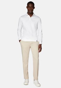 Balts garām piedurknēm polo krekls ar pogātu apkakli, kombinēts ar bēšiem biksēm un baltām sporta kurpēm, attēlots uz modeļa, kas stāv taisni.