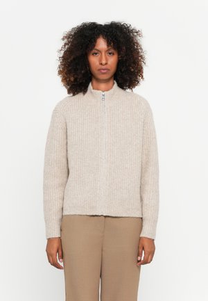Beige zip-up sweater with a ribbed texture, dropped shoulders, and a rounded collar, paired with light brown trousers.