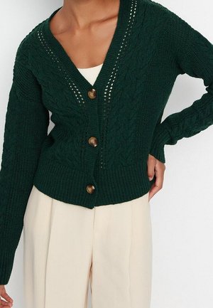 Cardigan vert foncé en maille torsadée avec trois boutons en bois, porté par-dessus un haut blanc, associé à un pantalon beige taille haute.