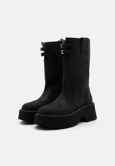 Botas negras hasta el tobillo con suela de plataforma gruesa, que cuentan con dos adornos de hebilla plateados y un material suave parecido al ante. Diseño simple y texturizado.