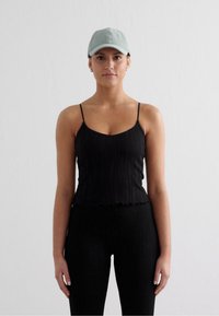 Svart ribbad tanktop med tunna remmar och en scalloped nedre kant, ihopparad med svarta leggings och en ljusgrön basebollkeps.