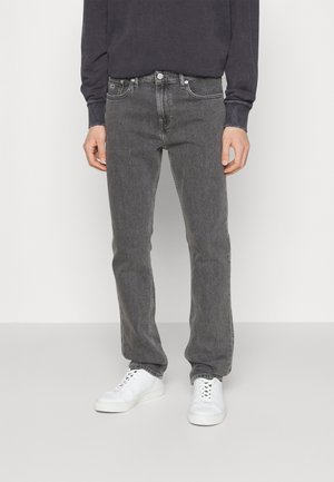 Homme portant un jean droit gris foncé, des baskets blanches et une chemise noire à manches longues, debout sur un sol gris contre un mur blanc.