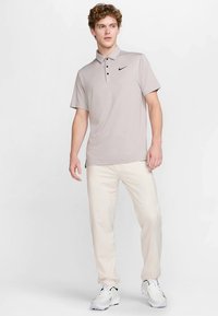 Ανοιχτό γκρι πόλο πουκάμισο με δύο κουμπιά και μαύρο λογότυπο Nike, σε συνδυασμό με μπεζ παντελόνια και λευκά αθλητικά παπούτσια. Λείο ύφασμα, casual σχεδιασμός.