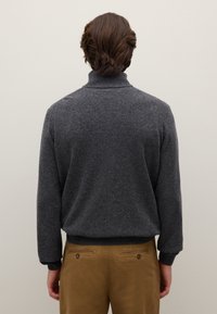 Maglione grigio scuro lavorato a maglia con collo alto e polsini a coste, indossato con pantaloni color cognac dotati di due tasche posteriori e dettagli con bottoni.