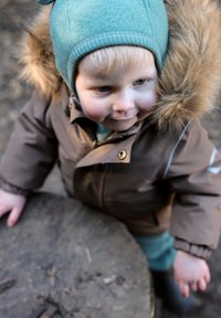 Brown puffy jacket with fur-lined hood, teal knit hat. Child resting hand on a wooden log, with light blond hair. Textured fabric details visible.