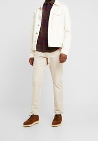 Giacca di denim bianca sopra una camicia a quadri scuri, chinos beige e scarpe in suede marrone con suola color crema. Outfit semplice e a strati.