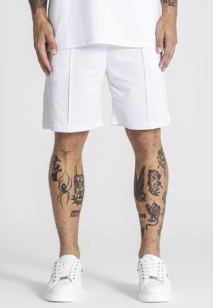 Homme portant un short blanc, des baskets blanches et une chemise blanche, montrant des jambes tatouées avec divers dessins à l'encre noire.