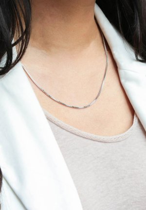 Zilveren ketting, plat en glad ontwerp, ongeveer 0,6 cm breed, gedragen op de huid met een licht shirt en een witte blazer.