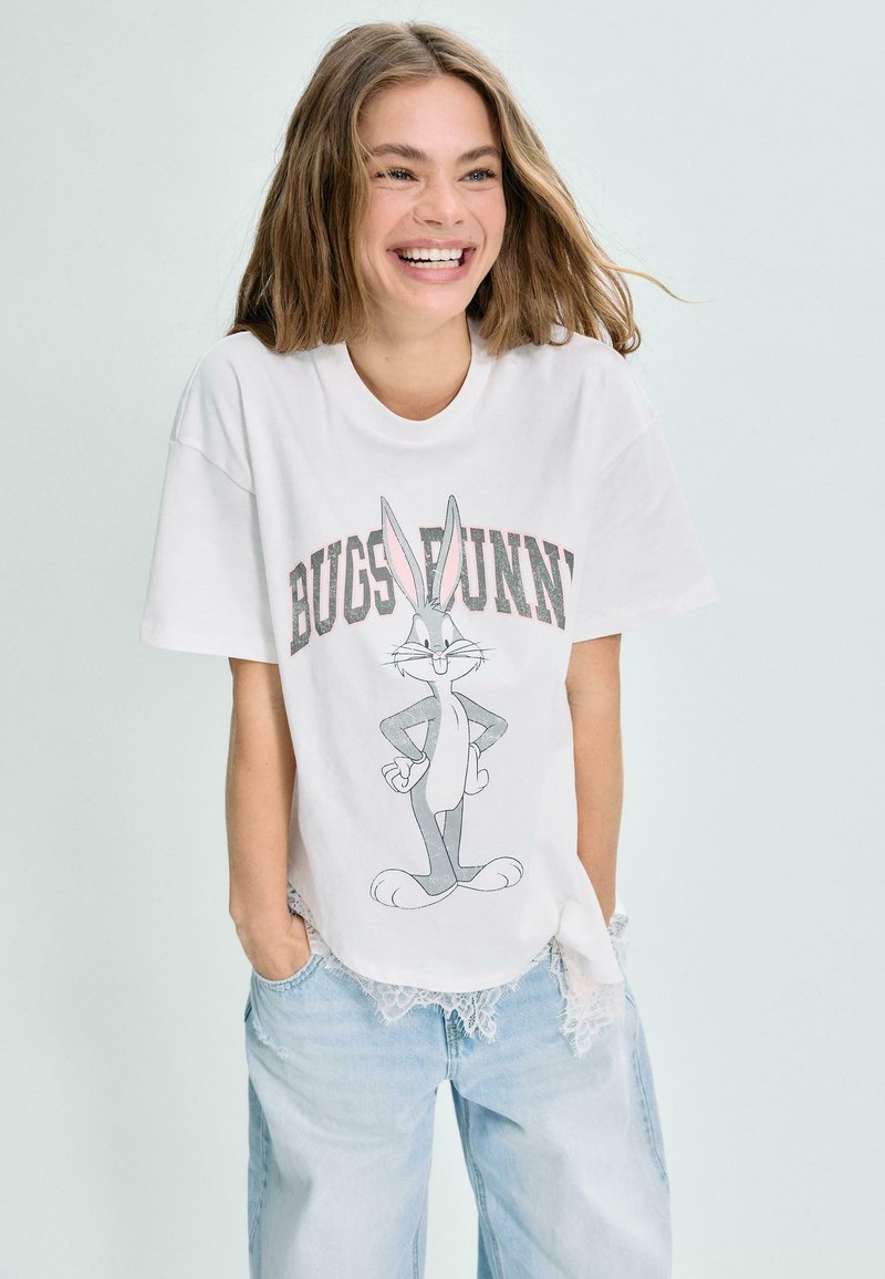 Jeune femme souriante, portant un t-shirt blanc avec un motif Bugs Bunny et un jean bleu clair, les mains dans les poches, sur un fond uni.