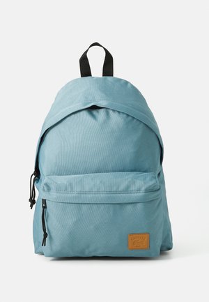 Mochila de tela azul con una parte superior redondeada, bolsillo frontal con cremallera, correas ajustables y una etiqueta de cuero marrón en el costado.