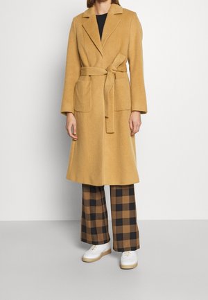 Vrouw met een lange, camelkleurige jas met ceintuur, zwart-bruin geruite broek en witte sneakers, staand tegen een effen achtergrond.