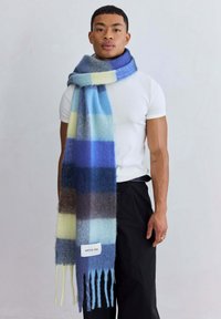 Geruite wollen sjaal met blauwe, gele en bruine tinten. Lang met rafelige uiteinden en een label met de tekst "ARCTIC FOX" aan de zijkant gestikt.