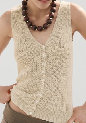 Veste sans manches beige en maille boutonnée, portée avec un grand collier de perles en bois foncé, les mains du modèle reposant sur les hanches.