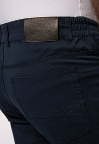 Marinblå stretchbyxor med ett strukturerat brunt läderbälte med en etikett som säger "BOSTON PARK." Har en bakficka och synliga sömnadsdetaljer.