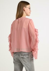 Helles rosa halbtransparente Bluse mit Rüschensäumen, mit einem Knopfverschluss am Rücken. Kombiniert mit grauen Jeans mit dekorativer Naht.