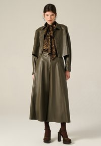 Giacca di pelle verde oliva con design corto e grandi tasche, abbinata a pantaloni larghi oliva e una sciarpa stampata marrone. Tacchi a piattaforma marroni.