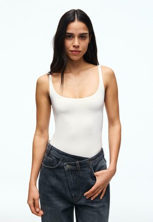 Woman with long dark hair wearing a white sleeveless top and loose dark jeans, standing with one hand touching her waistband.