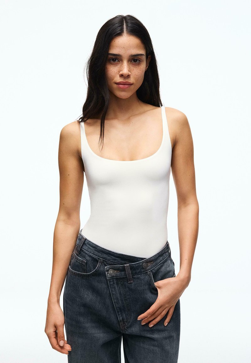 Femme aux longs cheveux foncés portant un haut blanc sans manches et un jean ample foncé, debout avec une main touchant sa taille.