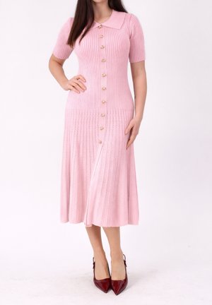 Vrouw die een roze geribbelde gebreide midijurk draagt met korte mouwen, een kraag, goudkleurige knopen aan de voorkant en bordeauxrode pumps met puntige neus.