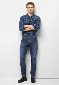 Blauwe geruite overhemden met lange mouwen, gecombineerd met donkerblauwe jeans en zwarte laarzen. Model poseert met de armen gekruist tegen een witte achtergrond.
