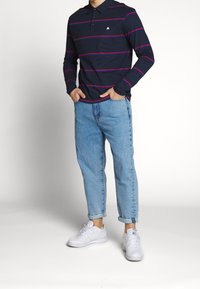 Laivaston pitkähiha polo, jossa on punaiset ja vaaleanpunaiset raidat, valkoinen logo rinnassa, yhdistettynä vaaleansinisiin, käännettyihin farkkuihin ja valkoisiin tennareihin.