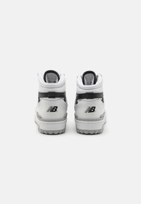 Bílé a šedé vysoké tenisky New Balance. Mají hladký kožený svršek, polstrovaný límec, texturovanou podrážku a značkovou logovou nálepku na patě.