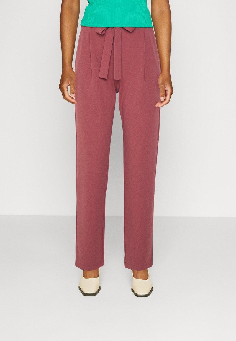 Femme portant un pantalon droit mauve taille haute avec une ceinture nouée, associé à des chaussures crème à bout carré et un top vert sans manches.