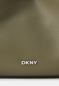 Geantă din piele verde măslinie cu suprafață texturată; are un logo DKNY din metal argintiu în partea de jos. Design simplu și funcțional.