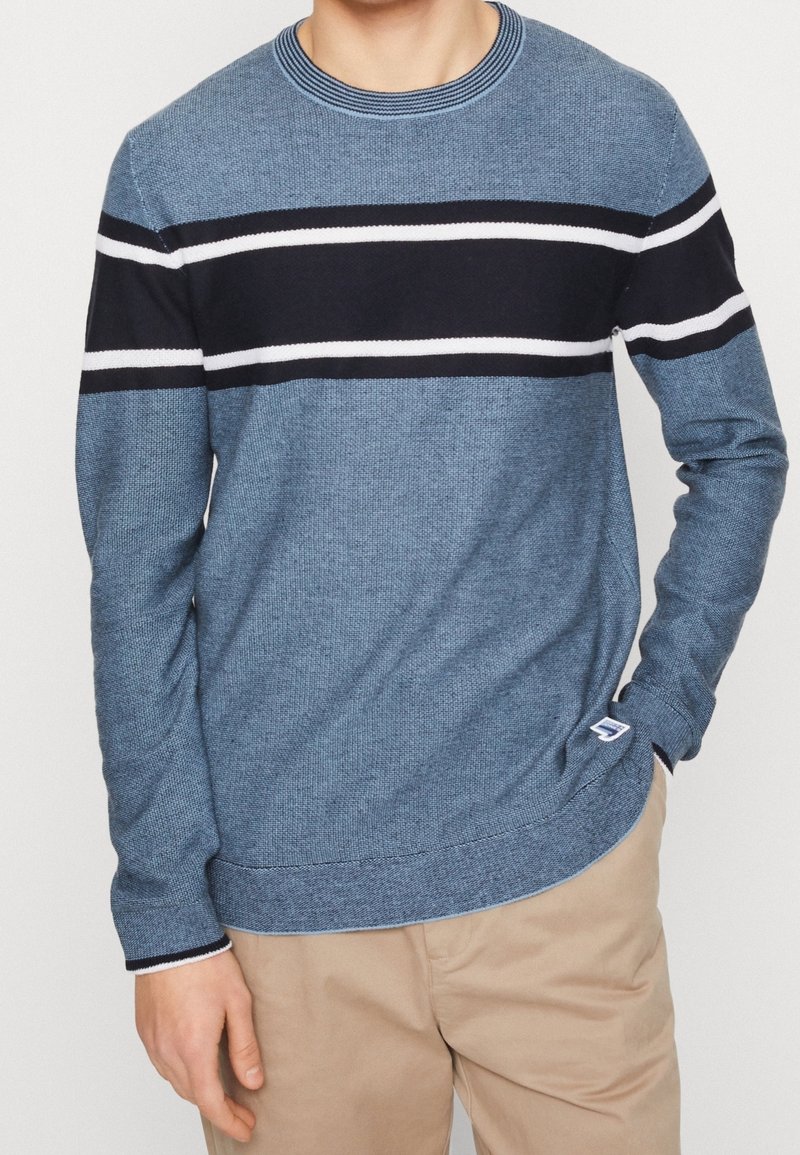 Mann trägt einen blauen Strickpullover mit schwarz-weißen horizontalen Streifen, kombiniert mit beigen Hosen, und steht mit einer Hand in der Tasche.