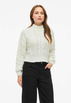 Pull en tricot blanc à col haut, avec des motifs en relief, des manches longues et une coupe courte, porté avec un pantalon noir taille haute.
