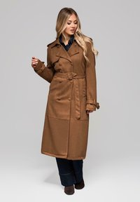 Abrigo trench coat marrón de doble botonadura con cinturón de lazo, que presenta hombreras y una textura semimate. Lleva puesto sobre pantalones vaqueros oscuros.
