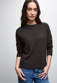 Dunkelbrauner Strickpullover mit rundem Halsausschnitt, gerippten Bündchen und Saum. Über blauen Jeans getragen, hat einen weichen Griff und eine lässige Passform.