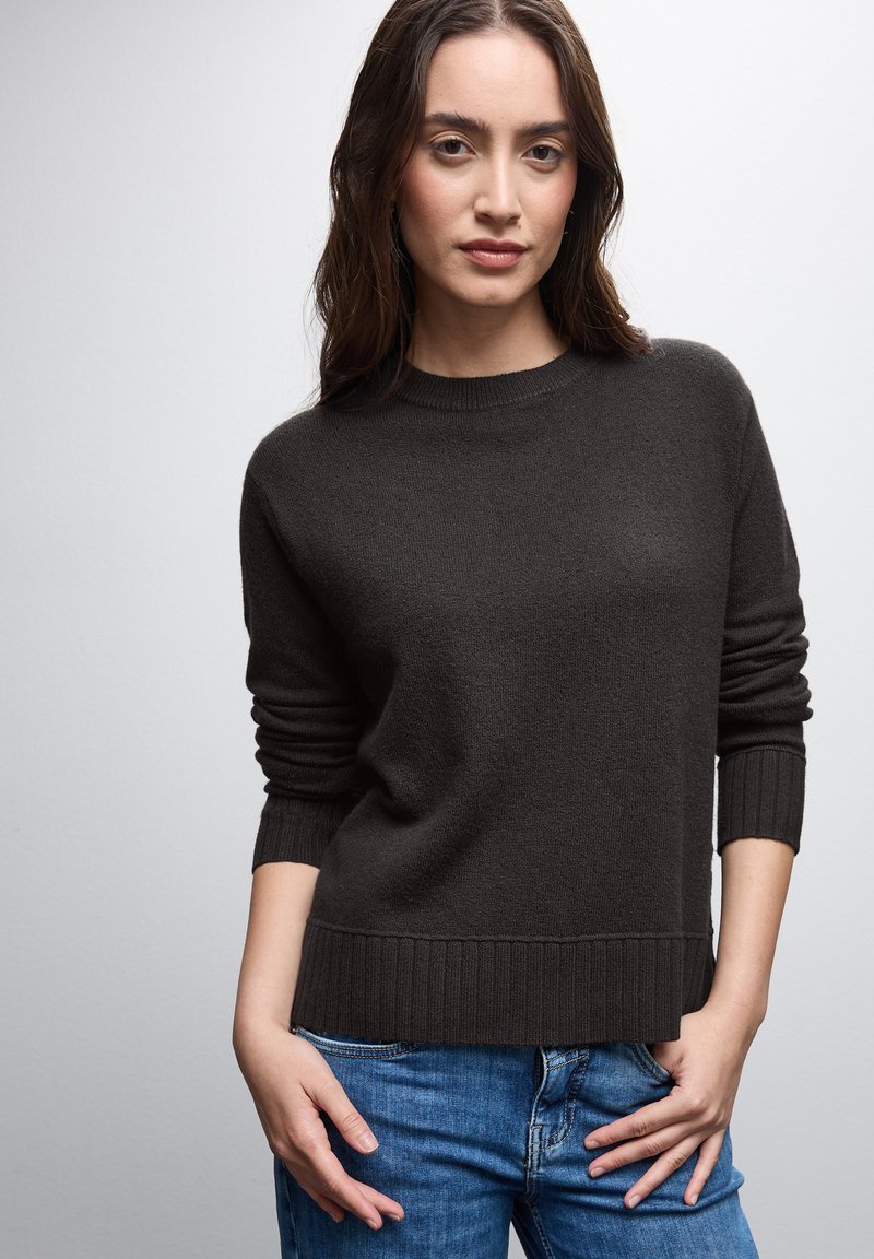 Dunkelbrauner Strickpullover mit rundem Halsausschnitt, gerippten Bündchen und Saum. Über blauen Jeans getragen, hat einen weichen Griff und eine lässige Passform.
