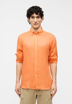 Man with curly dark hair wearing an orange long-sleeve button-up shirt with rolled sleeves and beige cargo pants.
