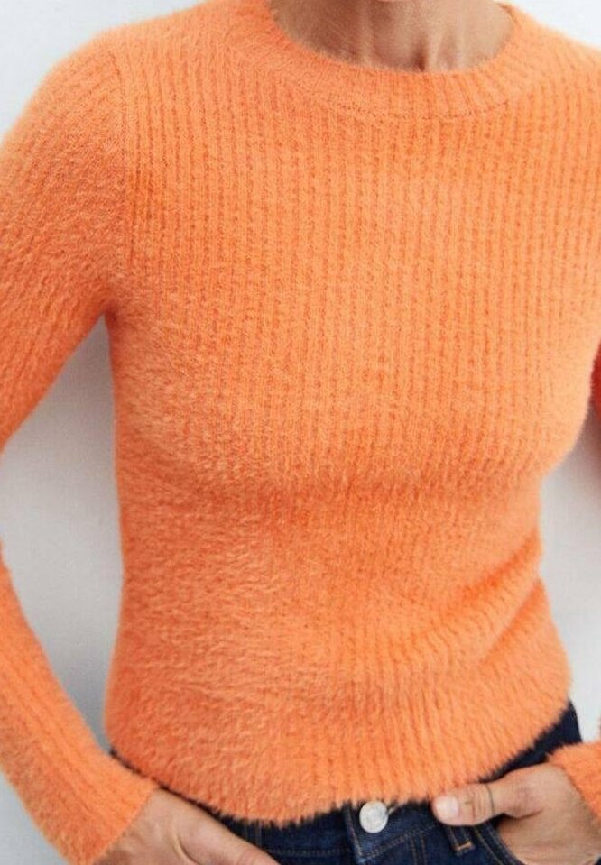 Gros plan d'une personne portant un pull orange ajusté et texturé et un jean bleu, les mains reposant près de la taille.