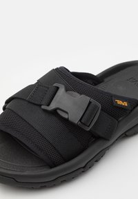 Svart sandal med texturerad remdesign, justerbart spänne och tjock gummisula. Anmärkningsvärd Teva-logotyp i orange.