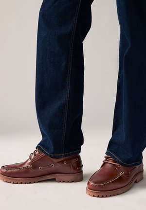 Zwei Beine mit dunkelblauen Jeans und braunen Leder-Schnür-Bootsschuhen mit Profilsohlen vor hellem Hintergrund.