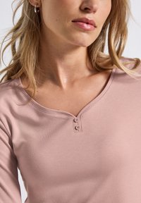 Femme portant un haut mauve clair avec un col en V et deux petits boutons, cheveux blonds lâches et boucles d'oreilles argentées en forme d'anneau visibles.