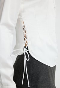 Witte blouse met lange mouwen en kanten details aan de zijkant, gedragen over donkere gestreepte broeken, waarbij een deel van de taille en arm van de drager zichtbaar is.