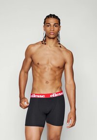 Schwarze Boxer-Briefs mit einem roten Elastikbund, auf dem der Markenname "ellesse" auffällig abgebildet ist. Der Stoff sieht glatt und eng anliegend aus.
