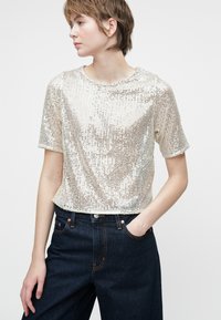 Top corto de manga corta hecho de brillantes lentejuelas plateadas, con un escote redondo y un dobladillo que llega a la cintura, combinado con jeans oscuros.