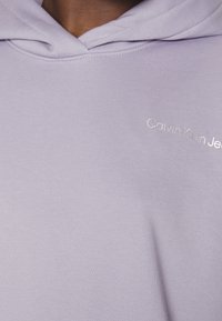 Liliowa bluza z kapturem wykonana z miękkiego materiału, z kieszenią z przodu, sznurkiem do regulacji kaptura oraz srebrnym logo "Calvin Klein Jeans" na klatce piersiowej.