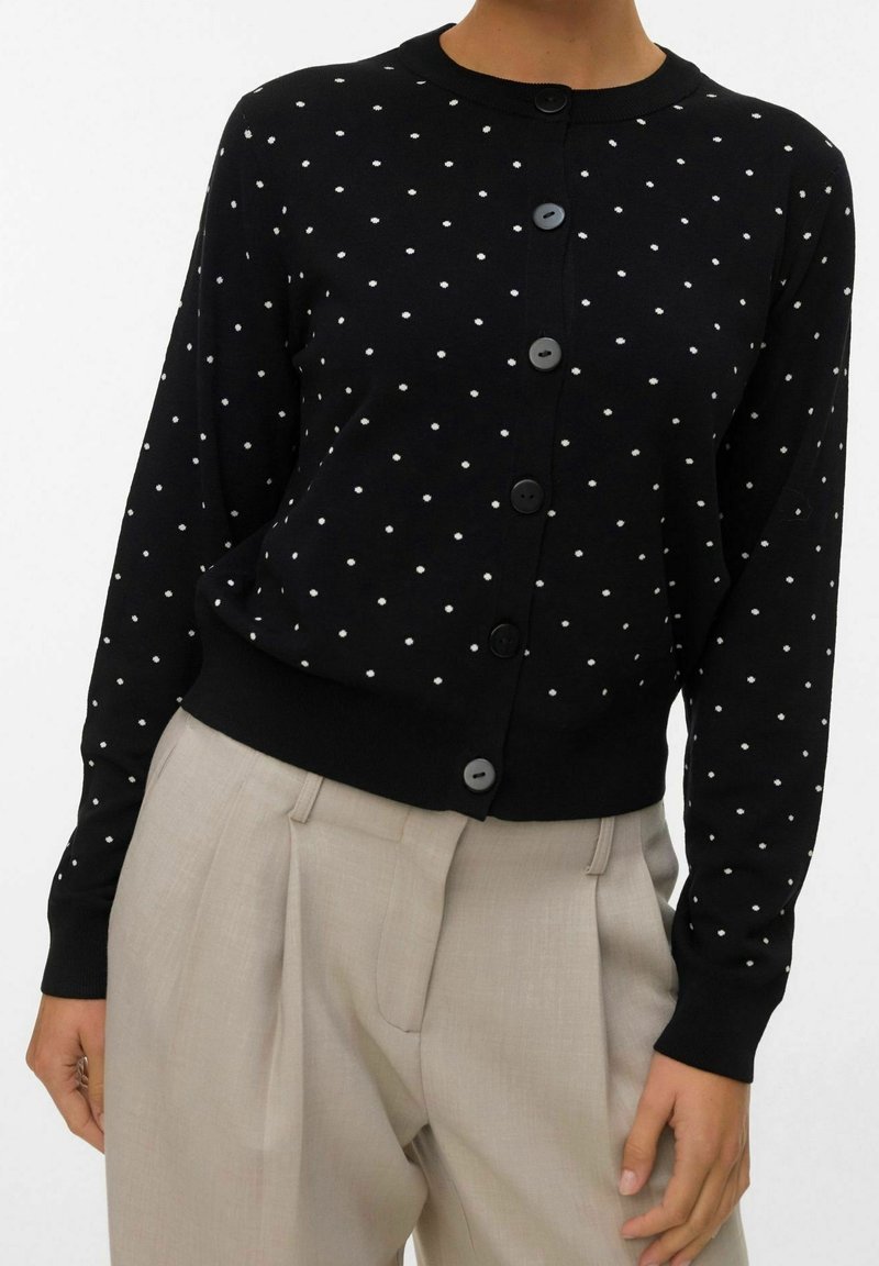 Cardigan noir à manches longues avec des pois blancs et des boutons noirs, associé à un pantalon beige plissé taille haute.