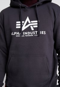 Sudadera gris oscura hecha de una mezcla de algodón con cordones ajustables. Cuenta con un bolsillo frontal y un logotipo blanco prominente en el pecho.