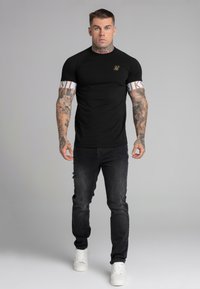 Melnā slim-fit T-kreklā ar baltajiem akcentiem un logo, kombinācijā ar nolietotām melnām džinsam un baltām kedām. Modelim ir tetovējumi un īsi mati.