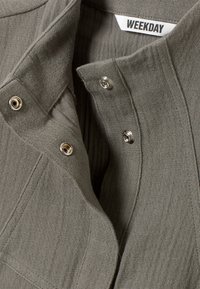 Chemise en tissu texturé vert olive avec fermetures à pression et col, présentant des rayures verticales subtiles et une étiquette de marque.