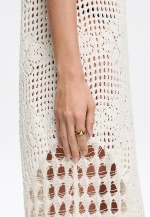 Abito in crochet crema con motivo a rete aperta. Mano appoggiata sul tessuto, che mostra un anello d'oro con un design astratto.
